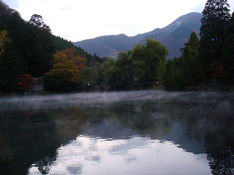 湯布院金鱗湖清晨霧景，湖面升起的霧氣與周邊樹木倒影形成如夢境般的景色